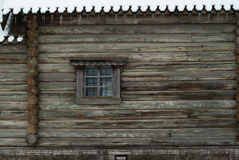 Заснеженная бревенчатая стена дома с обмёрзшим окном зимой