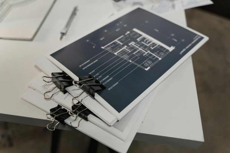 Стопка архитектурных планов с зажимами на белом столе перед финальной проверкой проекта