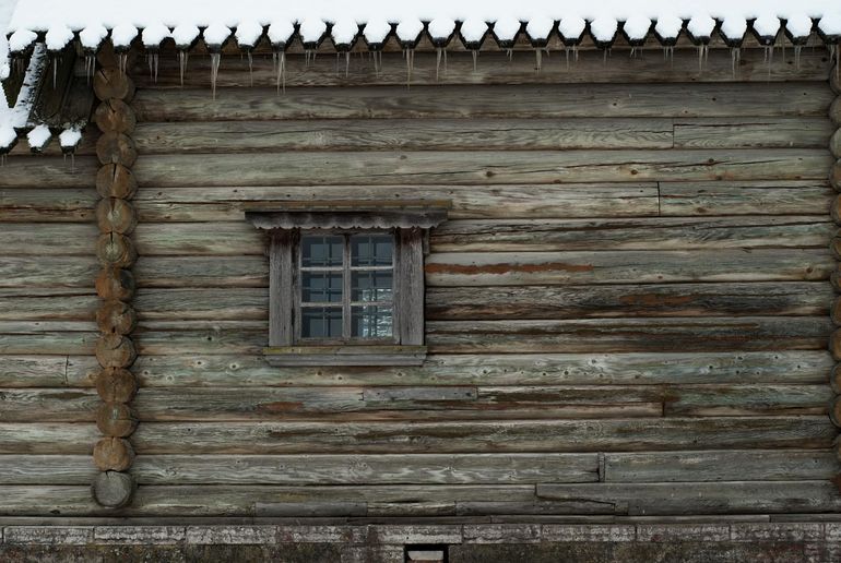 Заснеженная бревенчатая стена дома с обмёрзшим окном зимой