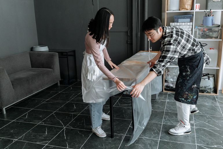 Двое укрывают стол плотной плёнкой перед малярными работами в студии